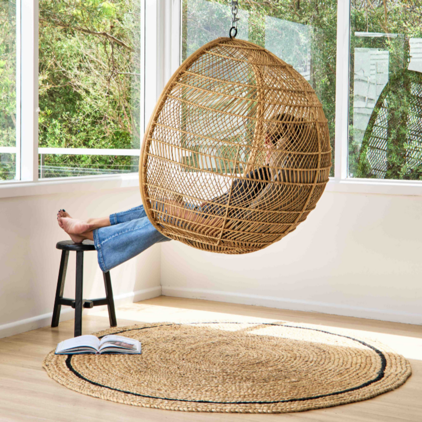 Round flatweave rug with graphite border sitting on light wooden floor in a room with large windows and greenery background and Person sitting in a hanging wicker chair