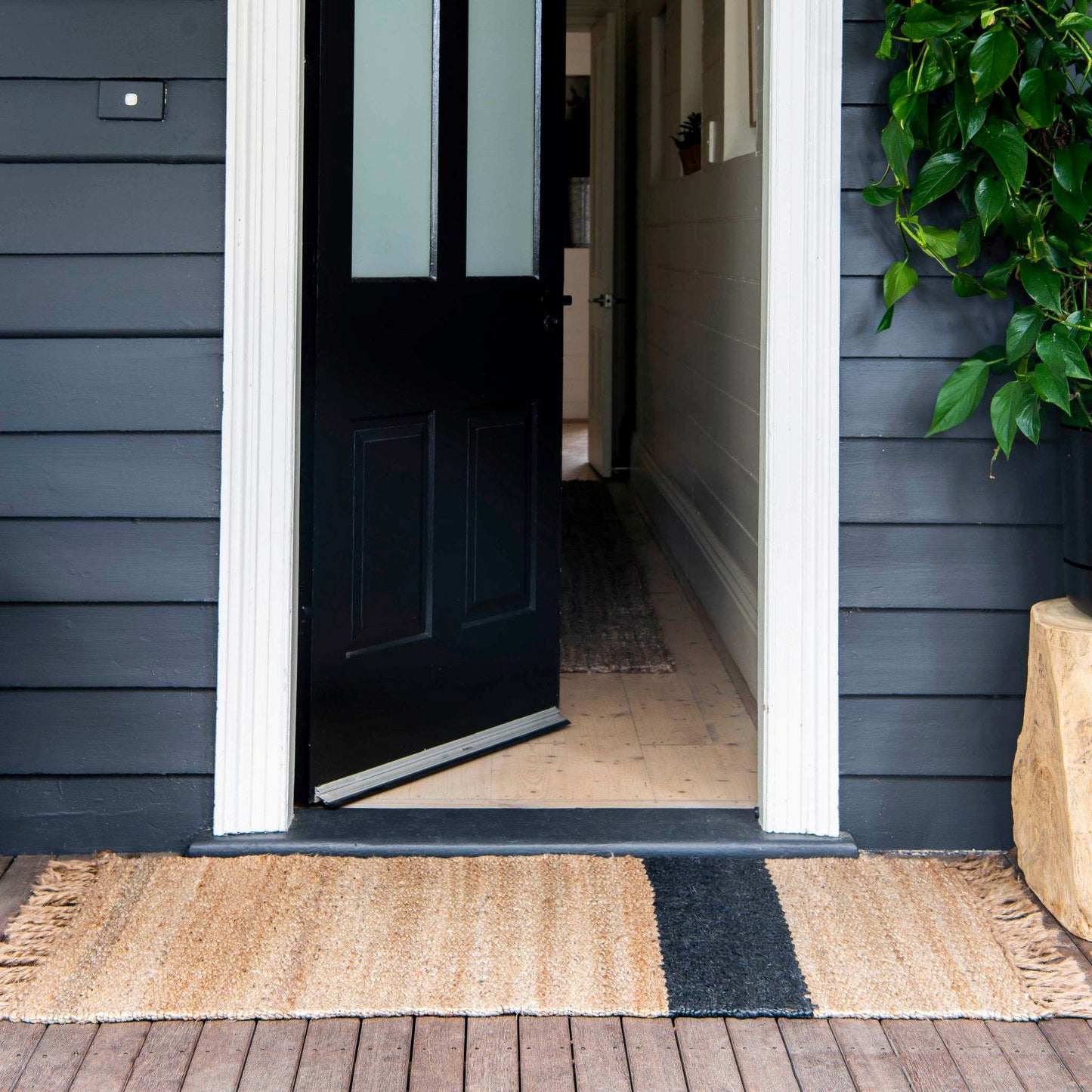 A natural jute Baxter rug with a graphite stripe, placed on a wooden deck in front of an open doorway.
