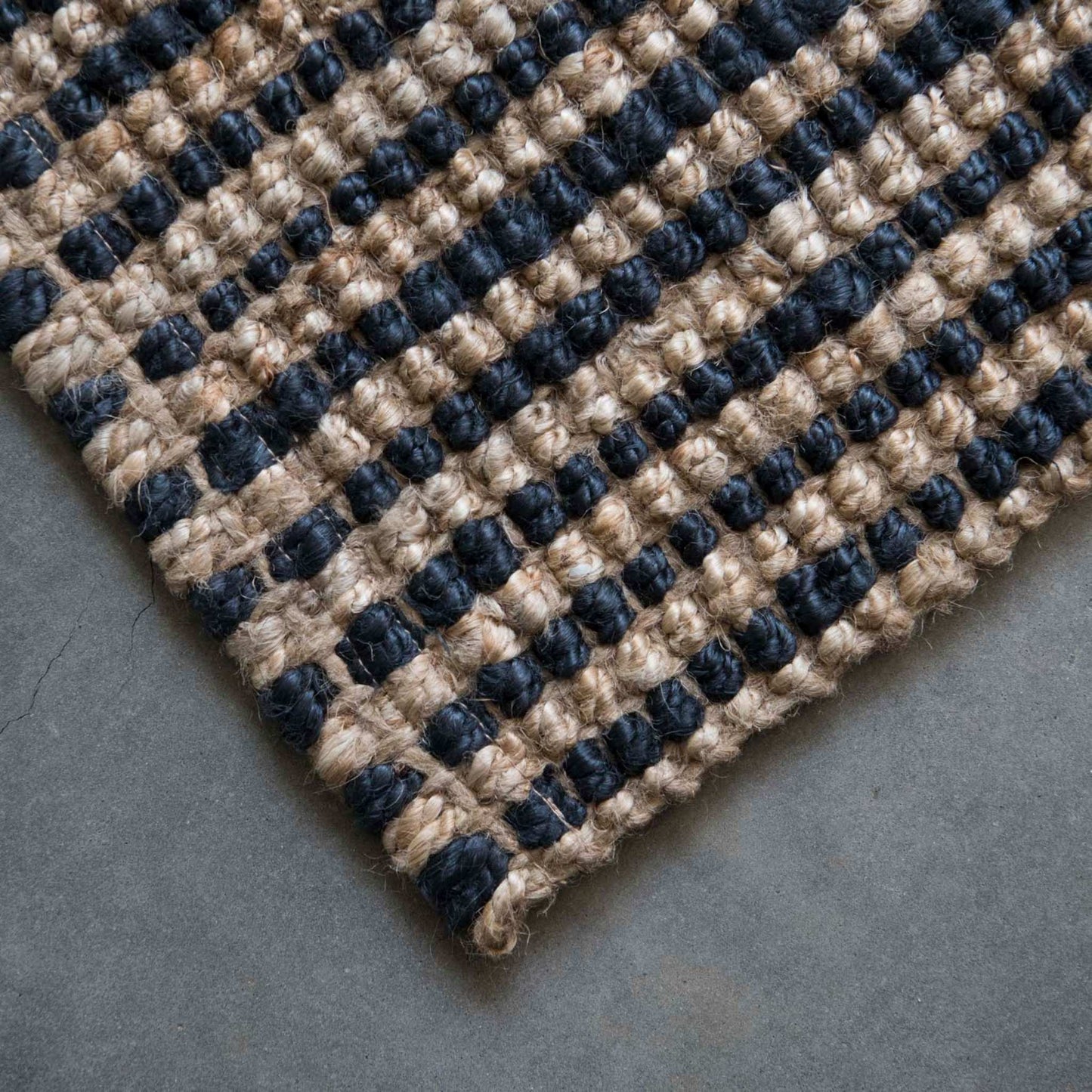 Close-up of the woven texture and graphite stripe on the Baxter jute rug with a brushed concrete background.