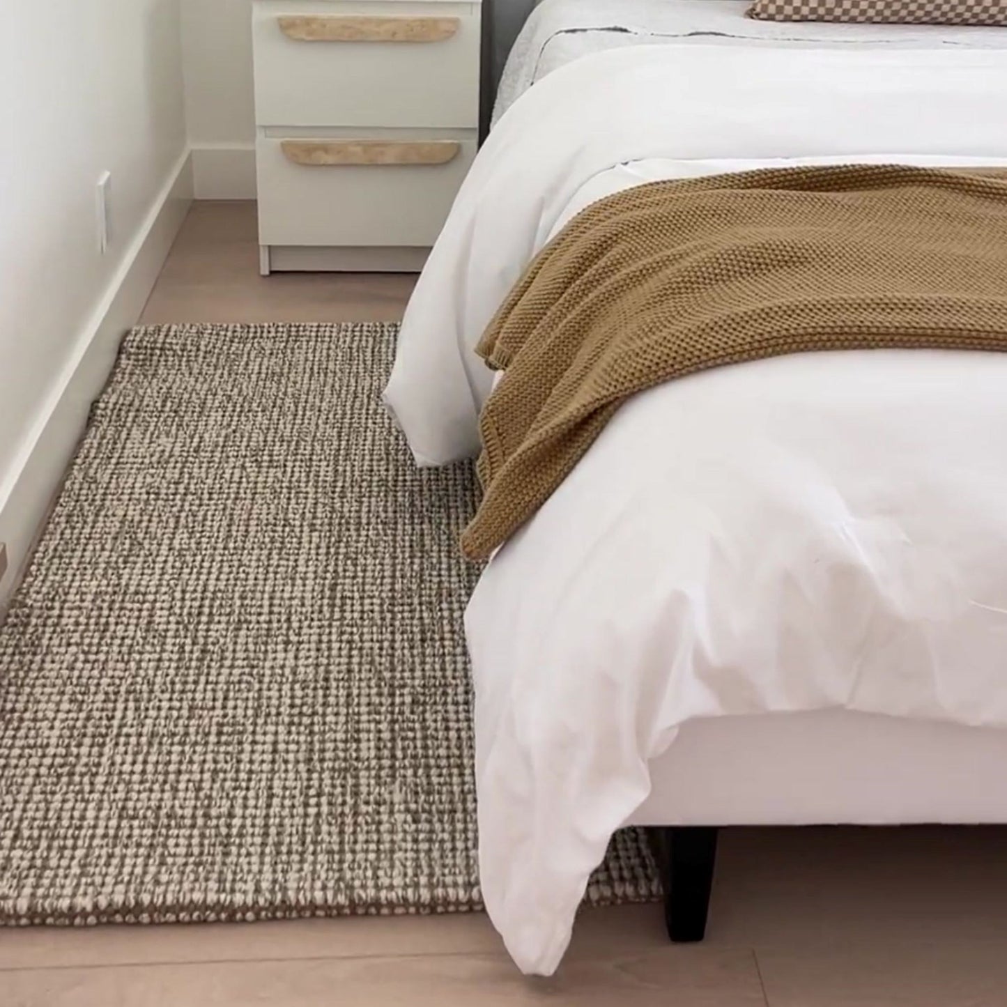 Carter handcrafted, grey and white heathered wool and jute bouclé small rug on a wooden floor next to a bed.