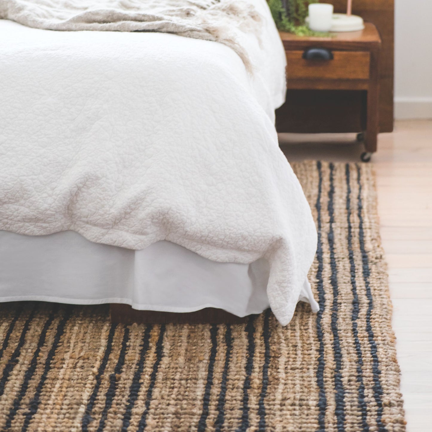 Smith natural bouclé jute area rug with graphite stripes on light wooden floor in a bedroom with white linen and dark timber furniture.
