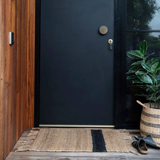 A natural jute Baxter rug with a graphite stripe, placed on a wooden deck in front of a black door.