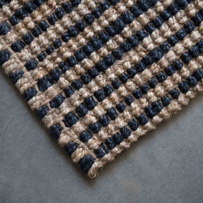 Close-up of the woven texture and graphite stripe on the Baxter jute rug with a brushed concrete background. 