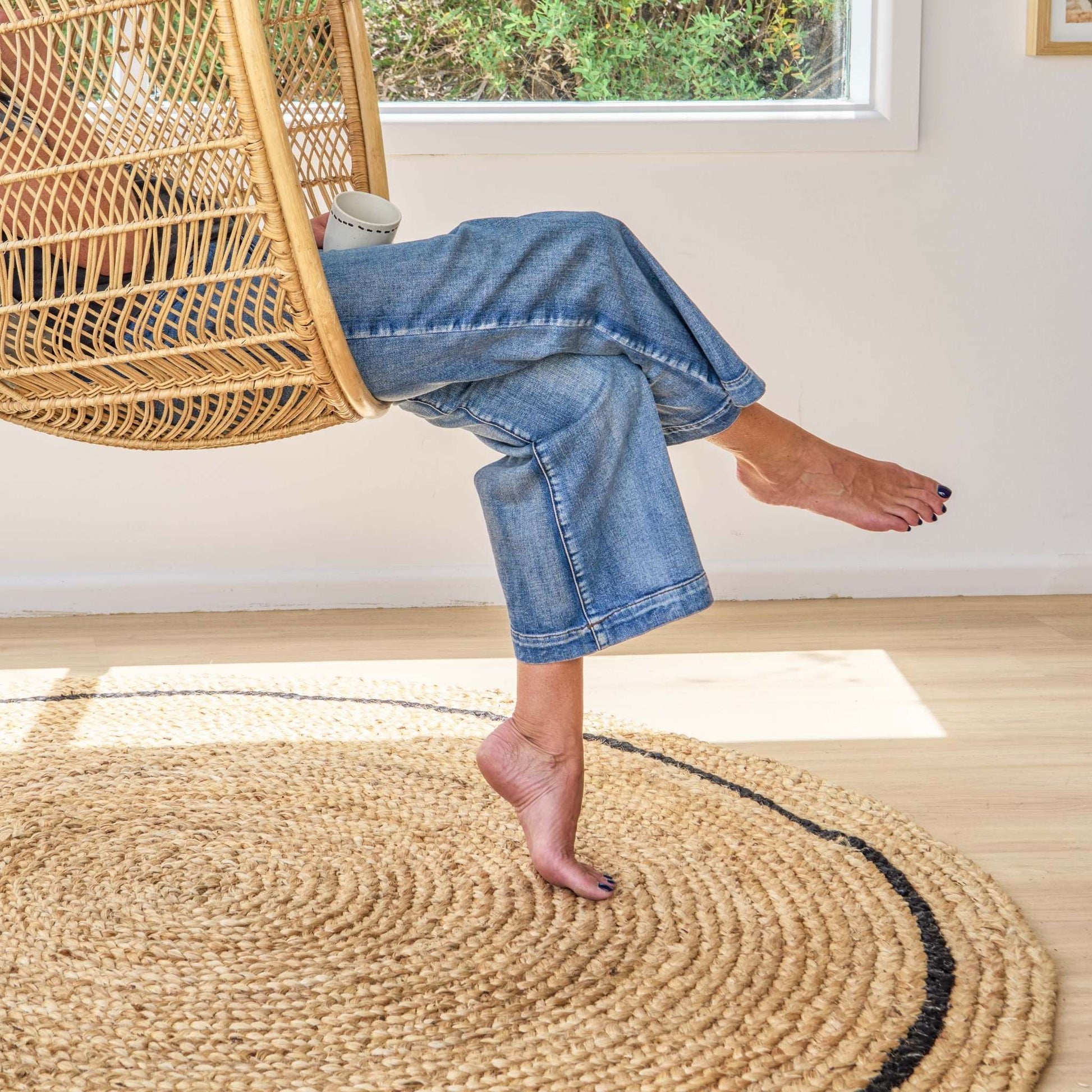 Cooper hand braided soft, jute, round rug with a graphite border on a wooden floor in sunlit room.