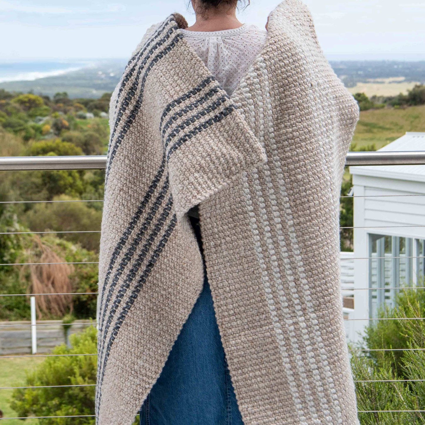 Macintosh flatweave rug, with natural and slate grey stripes made from 100% recycled PET, being held with the Franklin on a coastal balcony. 
