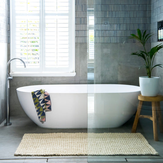 Vincent, whitewashed, chunky jute bouclé small rug on polished concrete in a coastal style bathroom with a white tub and timber stool.  