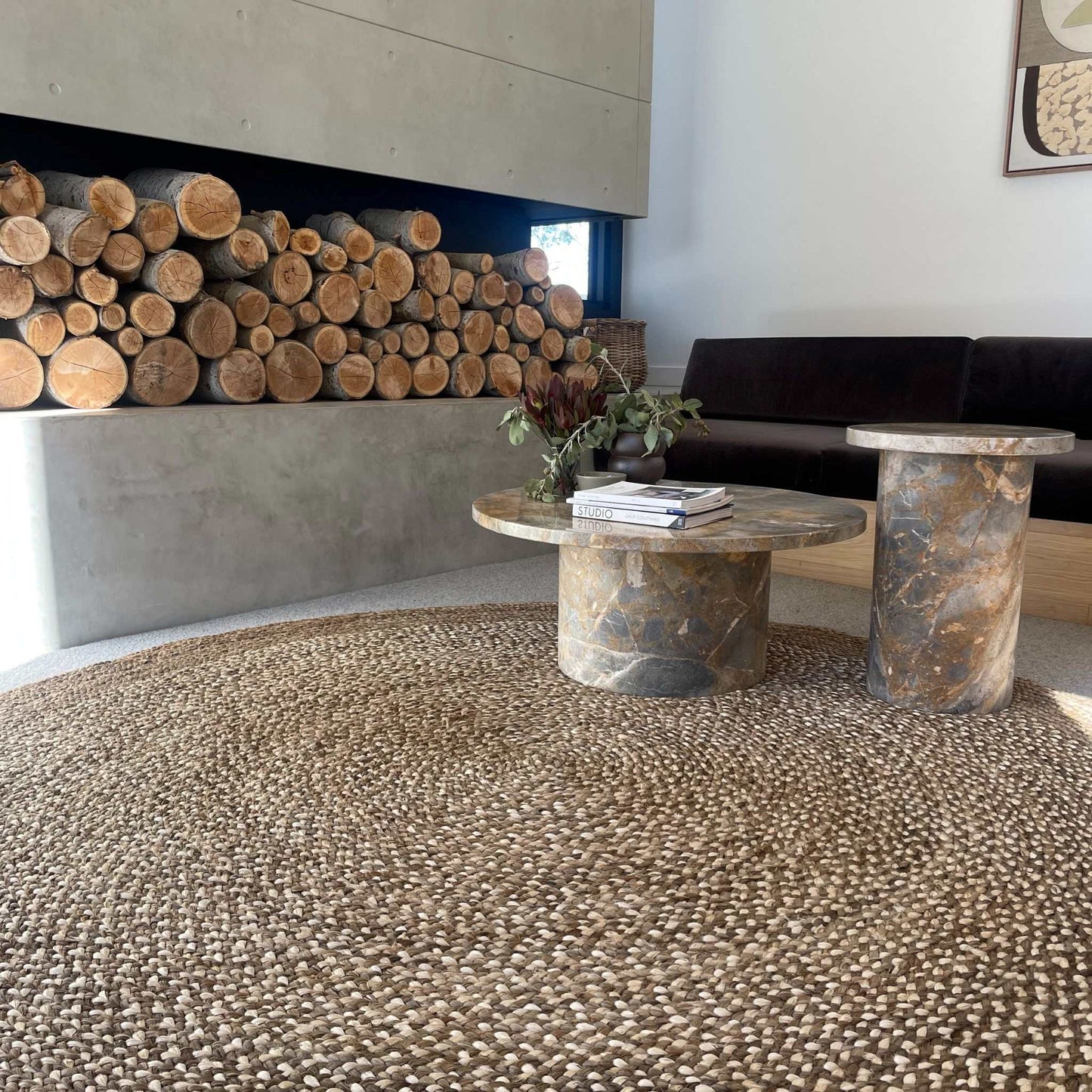Westmore hand-braided two toned jute round rug with a border sitting on polished concrete floor with a rustic wood pile. 