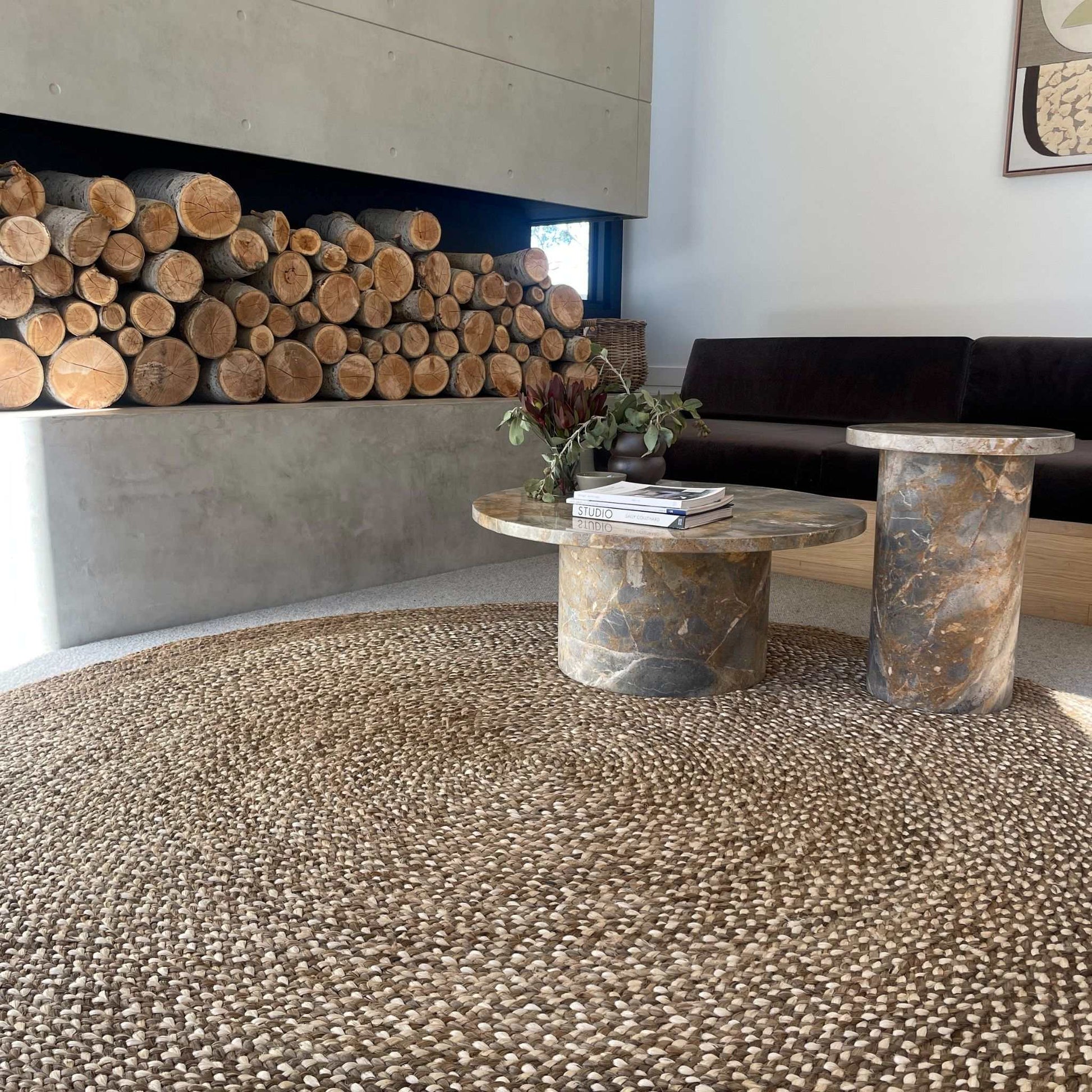 Westmore hand-braided two toned jute round rug with a border sitting on polished concrete floor with a rustic wood pile. 