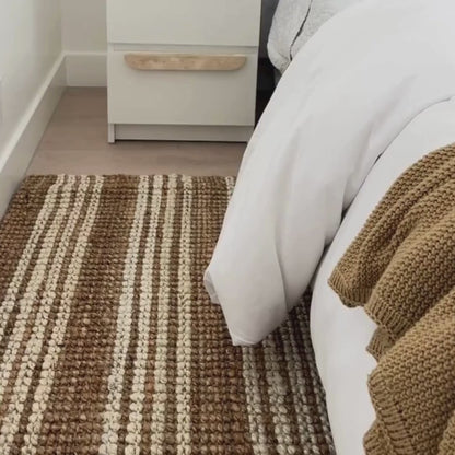 Close-up video of Osbourne natural and white striped jute rug with a chunky, bouclé weave next to a bed.